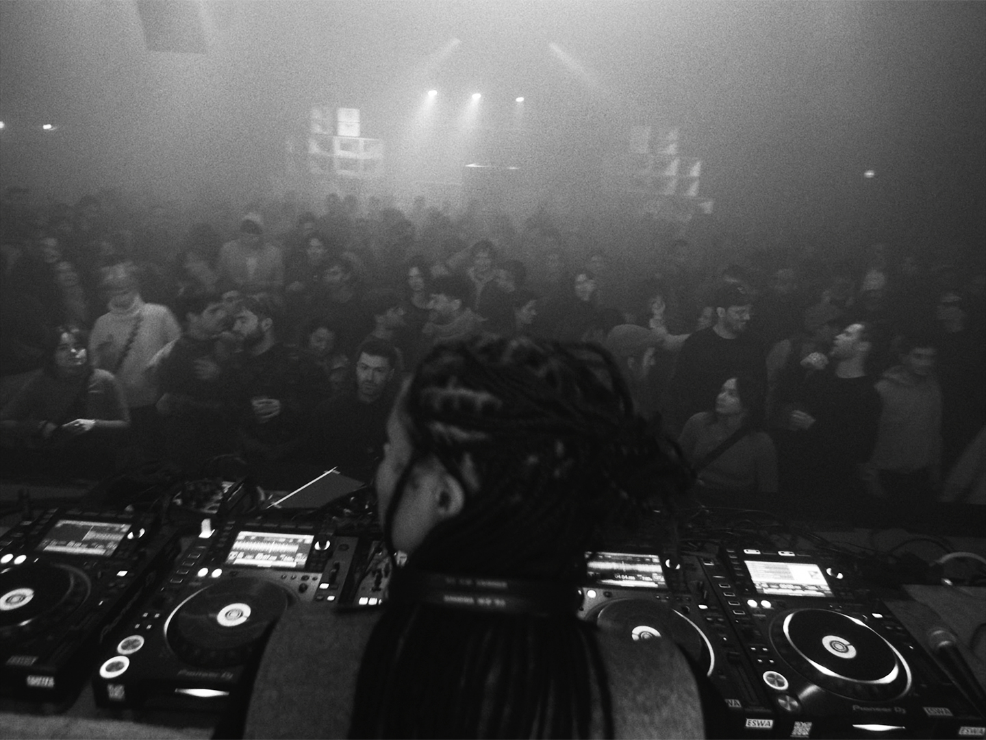 Black and white photo of Mia Koden behind a dj set, with a audience in front of here, and a big soundsystem in the back of the venue 